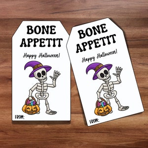 Könnte beinhalten: Zwei druckbare Geschenkanhänger für Halloween mit einem Skelett, das einen lila Hexenhut trägt und einen Kürbis-Eimer mit Süßigkeiten hält. Der Text auf den Anhängern lautet "Bone Appetit! Happy Halloween!" und "Von:"