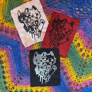 Puede incluir: Tres parches de tela con una ilustración en blanco y negro de dos gatos abrazándose. Los parches están sobre una superficie de crochet de colores. Los parches son en blanco y negro sobre un fondo beige, en blanco y negro sobre un fondo rojo y en blanco y negro sobre un fondo negro.