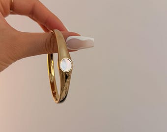 May include: Gold-tone bangle bracelet with a mother-of-pearl accent. The bracelet has a smooth, polished finish and a rounded shape. The mother-of-pearl accent is circular and set into the gold band. A simple, elegant piece of jewelry.