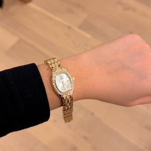 以下が含まれることがあります： 白い文字盤と小さな透明なクリスタルがちりばめられたベゼルが付いた、金色の腕時計。 時計には、テクスチャード加工のデザインが施された、お揃いのゴールドバンドが付いています。 時計は手首にあり、黒い袖が見えます。