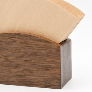 May include: A wooden coffee filter holder with a stack of brown paper coffee filters inside. The holder is made of dark wood with a natural grain pattern.