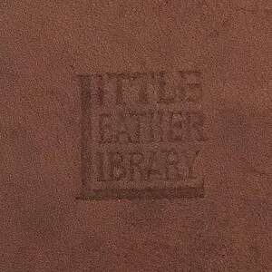 May include: Close-up of a textured, brown leather surface with the embossed text "Little Leather Library". The text is slightly darker than the surrounding leather, creating a subtle contrast. The image highlights the texture and color of the leather.