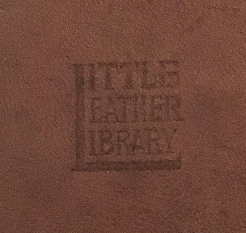 May include: Close-up of a textured, brown leather surface with the embossed text "Little Leather Library". The text is slightly darker than the surrounding leather, creating a subtle contrast. The image highlights the texture and color of the leather.