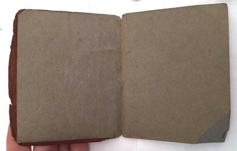 May include: An open, vintage book with aged, gray pages. The cover is a dark brown with visible wear and tear. The pages are slightly textured and show signs of age. The book is held open, revealing the interior pages.