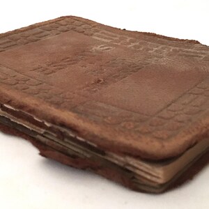 May include: A worn, brown leather-bound book with a rectangular shape. The cover has a textured pattern and the edges show signs of wear and tear. The pages are visible and appear aged, suggesting the book is antique.