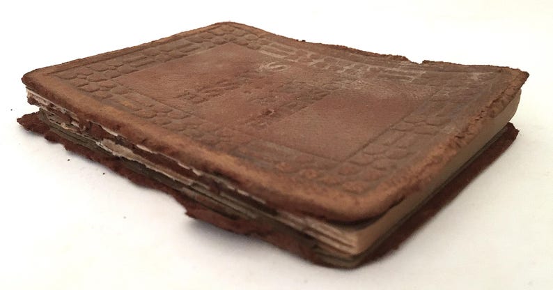 May include: A worn, brown leather-bound book with a rectangular shape. The cover has a textured pattern and the edges show signs of wear and tear. The pages are visible and appear aged, suggesting the book is antique.