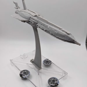 Puede incluir: Maqueta gris de nave espacial con detalles, montada sobre un soporte de plástico transparente. El modelo tiene un cuerpo largo y aerodinámico y está sostenido por un brazo gris curvado. Tres fichas circulares están colocadas en el soporte.