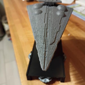 Puede incluir: Maqueta gris de una nave espacial Star Destroyer, con texturas superficiales detalladas y estructuras circulares prominentes. El modelo está montado sobre una base rectangular negra, que muestra un diseño triangular. Artículo de colección.