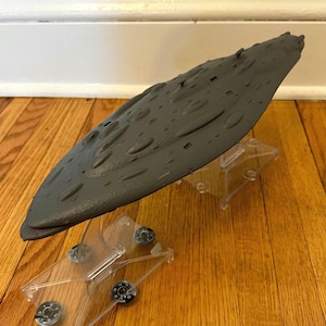 Puede incluir: Maqueta de nave espacial gris con superficie texturizada y detalles ovalados. El modelo está montado sobre dos soportes de plástico transparente. La nave espacial está sobre un suelo de madera y una pared blanca.