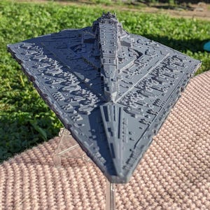 Puede incluir: Una maqueta gris de una nave estelar Star Destroyer, una nave espacial de ficción de una franquicia de ciencia ficción. El modelo está detallado con varias características de la superficie y está montado sobre un soporte transparente. La nave se exhibe al aire libre.