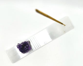 May include: A white selenite incense holder with a purple amethyst crystal on top. A wooden incense stick is placed in the holder.