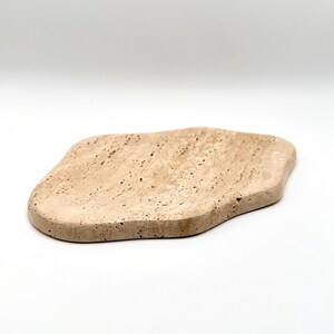 May include: A beige, organically shaped tray made of stone. The tray has a smooth surface with natural variations in color and texture. The edges are rounded, and the overall design is minimalist and modern. Perfect for serving or displaying items.