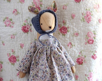 Könnte beinhalten: Eine handgefertigte Puppe mit einem Blumenkleid, einer blauen Strickmütze und braunen Stiefeln. Die Puppe hat ein rehähnliches Gesicht und steht auf einem floral gemusterten Stoff.