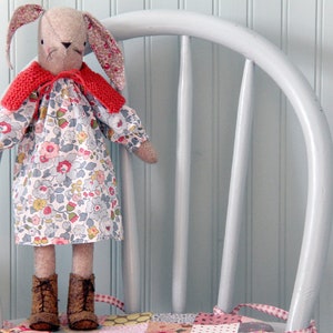 May include: A handmade bunny doll with a floral print dress, a red knitted collar, and brown boots. The bunny has long ears with a pink floral pattern and is standing on a white chair.