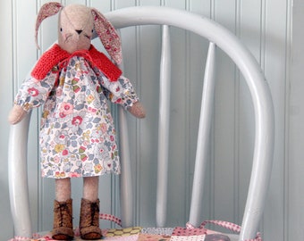 Könnte beinhalten: Handgemachte Hasenpuppe mit geblümtem Kleid, rotem Strickkragen und braunen Stiefeln. Der Hase hat lange Ohren mit einem rosa Blumenmuster. Die Puppe steht auf einem weißen Stuhl.