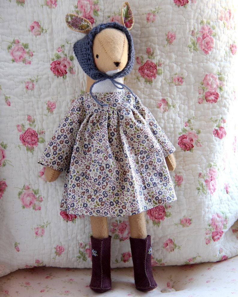 May include: A handmade doll with a floral dress, blue knitted hat, and brown boots. The doll has a deer-like face and is standing on a floral patterned fabric.