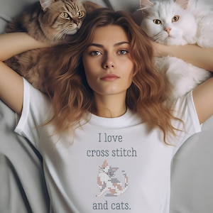 May include: A woman wearing a white t-shirt with the text "I love cross stitch and cats." and a cross-stitch design of a cat on the front. She is lying on a bed with two cats, one white and one brown tabby.