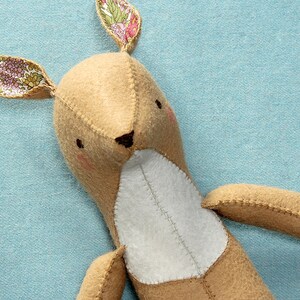 May include: A brown felt stuffed animal with a white belly and floral fabric ears. The animal is lying on a light blue surface.