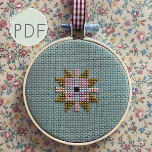 May include: A teal cross-stitch embroidery hoop with a yellow, pink, and brown geometric star design. The hoop is hanging from a red and white striped ribbon.