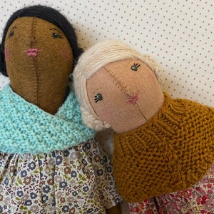 K&ouml;nnte beinhalten: Zwei handgefertigte Filzpuppen, die gestrickte Schals und Blumenkleider tragen. Die Puppe links hat braune Haut und schwarze Haare, w&auml;hrend die Puppe rechts helle Haut und blonde Haare hat.