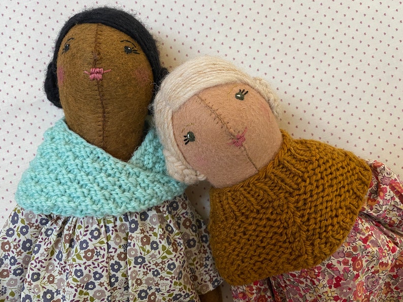 K&ouml;nnte beinhalten: Zwei handgefertigte Filzpuppen, die gestrickte Schals und Blumenkleider tragen. Die Puppe links hat braune Haut und schwarze Haare, w&auml;hrend die Puppe rechts helle Haut und blonde Haare hat.