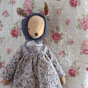 May include: A handmade doll with a blue knitted hat and a floral dress. The doll is sitting on a white quilted fabric with a floral pattern.