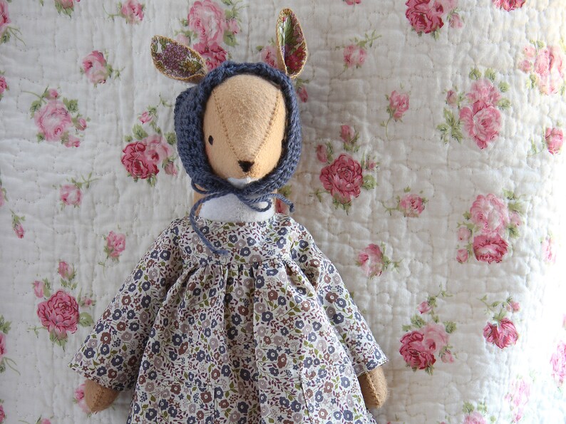 May include: A handmade doll with a blue knitted hat and a floral dress. The doll is sitting on a white quilted fabric with a floral pattern.