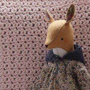 May include: A handmade plush fox toy wearing a blue hooded dress with a floral pattern. The fox has brown eyes and a long, pointed nose. The toy is sitting on a pink crocheted blanket.