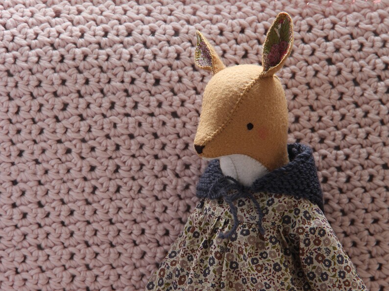 May include: A handmade plush fox toy wearing a blue hooded dress with a floral pattern. The fox has brown eyes and a long, pointed nose. The toy is sitting on a pink crocheted blanket.