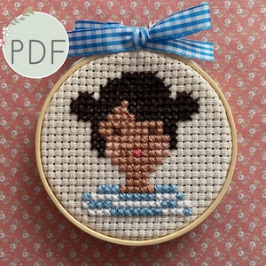 May include: A cross-stitch embroidery of a person with brown hair and skin, wearing a blue and white striped shirt, framed in a wooden hoop.