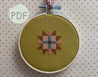 May include: A cross-stitch pattern with a geometric design in shades of brown, pink, and green, stitched on a mustard yellow fabric. The fabric is mounted in a wooden embroidery hoop.