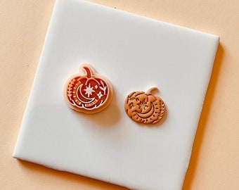May include: Two small orange pumpkin-shaped earrings with white and gold details. The earrings are on a white tile surface.