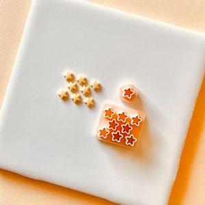 May include: A white ceramic tile with two pink plastic star-shaped cookie cutters. One cutter is a single star, the other is a grid of stars. There are also small yellow star-shaped sprinkles on the tile.