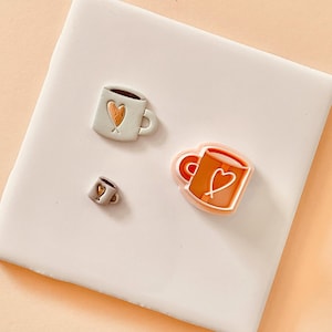 May include: Three ceramic coffee mugs with heart designs. The mugs are in different sizes and colours, including white, brown, and orange.