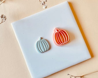 May include: Two pumpkin-shaped cookie cutters, one light blue and one pink, on a white ceramic tile.