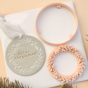 May include: A light gray Christmas ornament with the text "first christmas" in copper-colored letters. The ornament is round with a white floral design around the edge. A white ribbon is attached for hanging. A light pink wreath and a cookie cutter are also in the image.