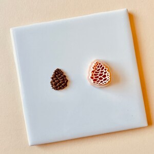 May include: Two pine cone shaped polymer clay ornaments. One is brown and the other is pink with a detailed design.