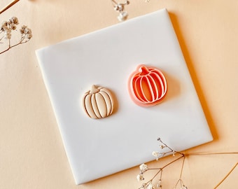 May include: Two pumpkin-shaped items sit on a white square against a peach background. One is a beige pumpkin-shaped cookie, and the other is a pink pumpkin-shaped cookie cutter. The items are surrounded by delicate white flowers.