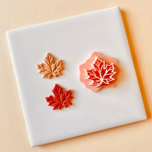 May include: Three polymer clay maple leaf shapes in various shades of brown and red. The leaves are on a white tile surface.