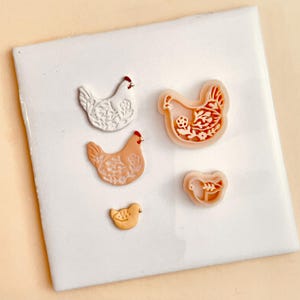 May include: Four ceramic chicken cookie cutters, one white, two peach, and one yellow. The cutters are arranged on a white surface.