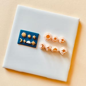 May include: A set of 10 small, pink plastic cookie cutters in the shapes of a sun, star, crescent moon, cloud, and a few other shapes. The cutters are arranged on a white surface next to a blue card with gold-colored cookie cutters in the same shapes.