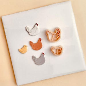 May include: A set of six cookie cutters shaped like chickens. The cutters are made of white plastic and feature different chicken designs, including a chick, a hen, and a rooster.