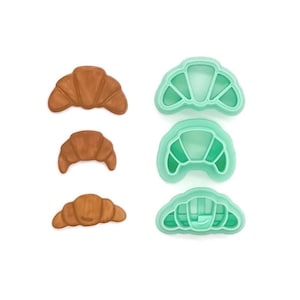 May include: The image shows three baked croissants and three teal-colored croissant-shaped cookie cutters. The croissants are golden brown, and the cookie cutters are made of plastic. The items are arranged on a white background.