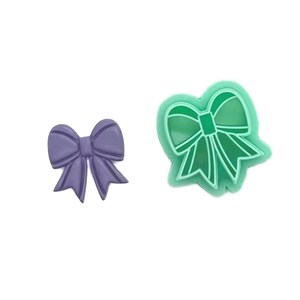 May include: Two bow-shaped items. One is a solid, lavender-coloured bow. The other is a green bow-shaped cookie cutter. The cookie cutter has a green outline and a hollow centre.