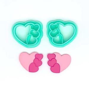 May include: Two sets of heart-shaped cookie cutters and two heart-shaped fondant decorations. The cookie cutters are a bright teal color, and the fondant decorations are pink and dark pink. The design features a large heart with three smaller hearts.