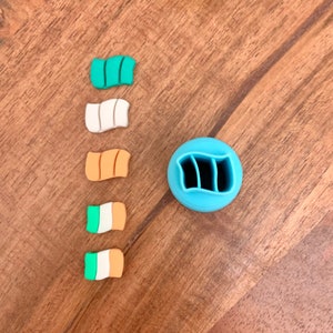 Puede incluir: Una colección de coloridos dijes de arcilla polimérica con forma de bandera y un cortador azul a juego. Los dijes presentan diseños en verde, blanco y naranja, que se asemejan a banderas. Los artículos están dispuestos sobre una superficie de madera.