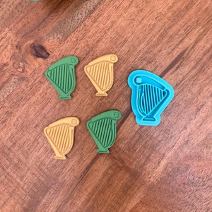 May include: A collection of harp-shaped cookie cutters and cookies in shades of green, yellow, and blue. The harp-shaped objects are arranged on a wooden surface, with a green leaf in the upper left corner.