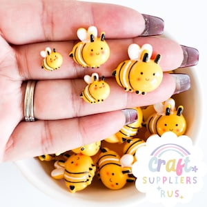 以下が含まれることがあります： 黄色と黒のハチの形をしたクラフト装飾品のコレクション。ハチは白い羽と笑顔の顔をしています。手で持っているものもあれば、白いボウルに入っているものもあります。Craft Suppliers Rusのロゴが見えます。