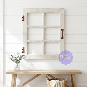 Op de afbeelding: Een verweerd wit houten raamkozijn met zes ruiten, donkerbruine scharnieren en een handvat. Het frame is gemonteerd op een witte lambrisering muur boven een houten bank met een glazen vaas en een rieten mand.