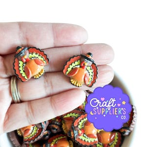 May include: Two small turkey-shaped flatback cabochons featuring brown bodies, red wattles, and colorful yellow, orange, and red tail feathers. Each turkey wears a small black hat. The items are shown in a pile with a purple Craft Suppliers Co. logo.